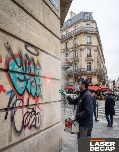 Home Opérateur retirant des graffitis sur un mur en pierre à Paris à l’aide d’un laser de décapage, équipé d’un masque et d’un casque de protection.